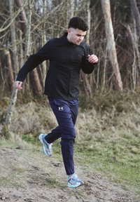 Black zip-up running jacket, navy athletic trousers, and grey-blue running shoes. The runner is in motion against a natural terrain backdrop.