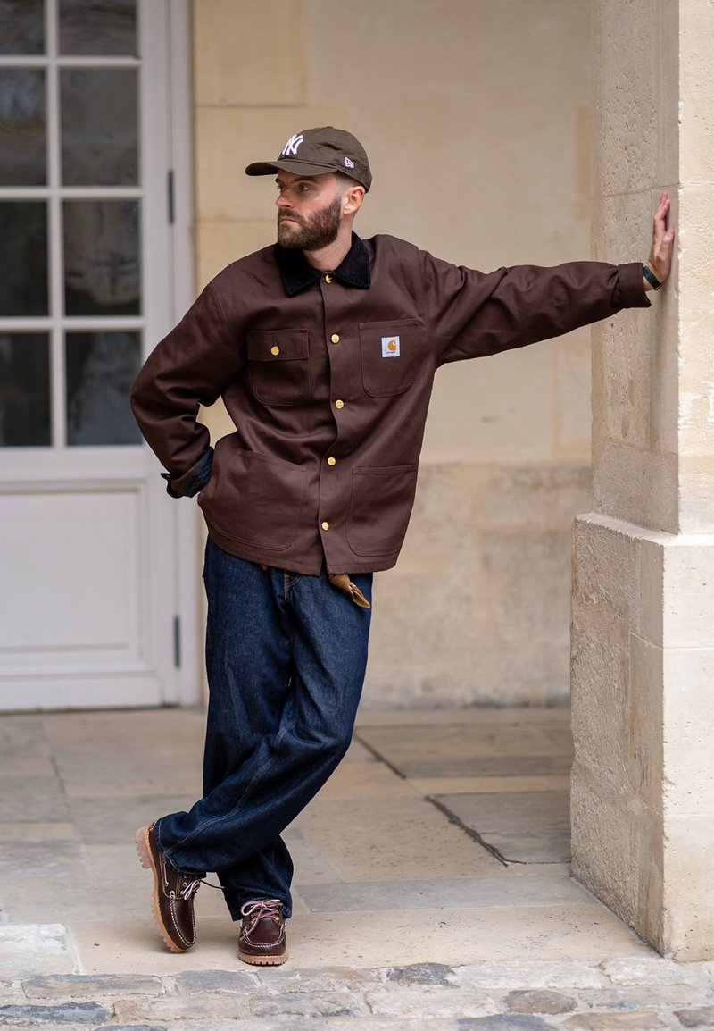 Jacket de trabalho castanho com colarinho preto, botões dourados e dois bolsos frontais; combinado com calças de ganga azuis e sapatos castanhos com atacadores.