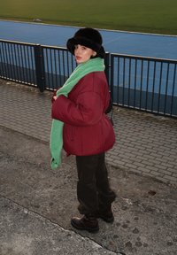 Μαρον πούφερ μπουφάν, πράσινο κασκόλ, μαύρο γούνινο καπέλο, καφέ παντελόνι και σκούρες μπότες. Εξωτερικός χώρος με διαδρομή τρεξίματος στο βάθος.