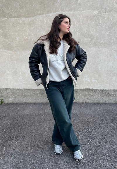 Chaqueta de cuero negra con forro de borrego, sudadera gris con texto, pantalones verde oscuro y zapatillas grises. Conjunto urbano casual.