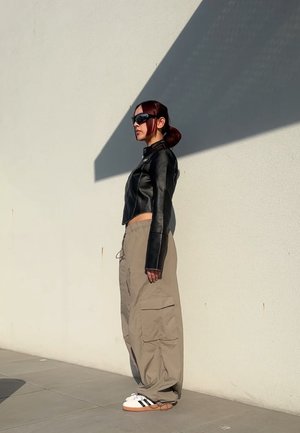 Black leather cropped jacket, beige cargo pants with side pockets, and white sneakers with black stripes. Sunglasses and a smooth hairstyle.