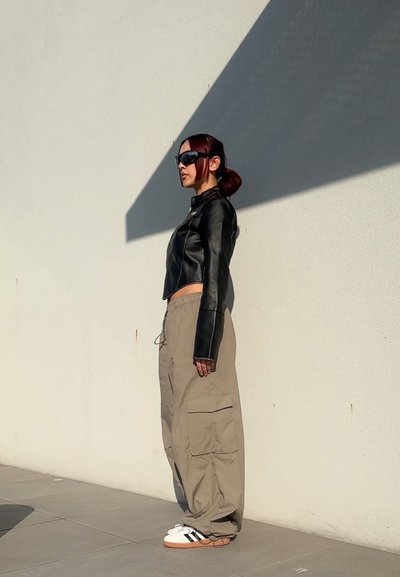 Chaqueta de cuero negra corta, pantalones cargo beige con bolsillos laterales y zapatillas blancas con rayas negras. Gafas de sol y un peinado liso.