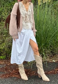 Cárdigan de punto rosa claro, blusa blanca con volantes, falda blanca con abertura frontal y botas de ante sintético color beige hasta la rodilla con detalle texturizado.
