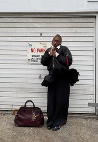 Geacă bomber neagră cu guler alb, pantaloni negri largi și o geantă vișinie cu mâner din lemn, decorată cu accesorii de tip breloc.