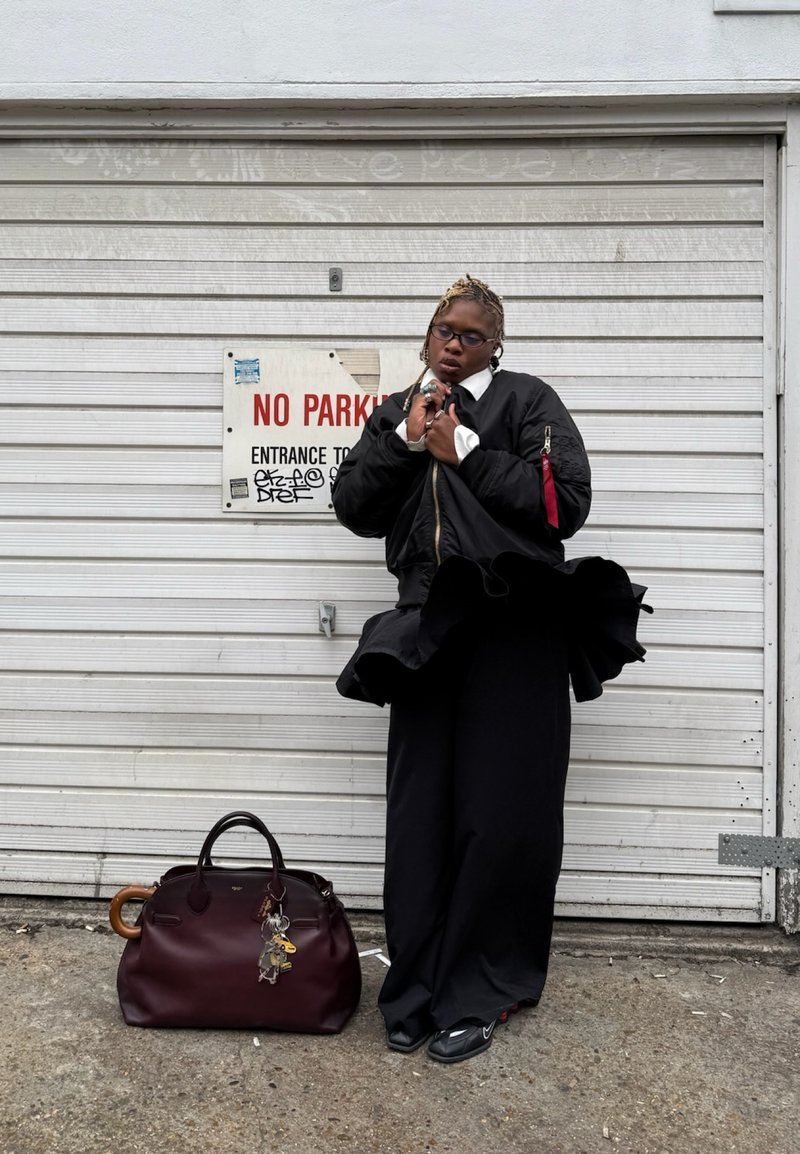 Svart bomberjakke med hvit krage, svarte vide bukser, og en burgunder veske med håndtak i tre, pyntet med nøkleringsanheng.