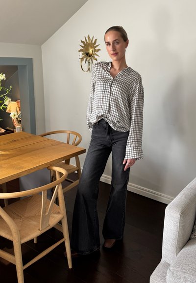 Camisa de cuadros en blanco y gris con mangas anchas, combinada con jeans de tiro alto negros de campana. Mesa de madera y silla beige de fondo.