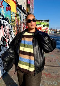 Giacca di pelle nera su un maglione peloso a righe multicolori (marrone, verde, blu, crema). Tiene una borsa nera texturizzata su uno sfondo di graffiti.