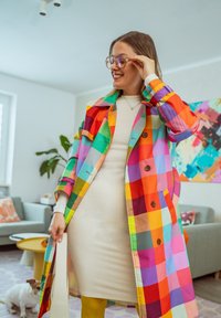 Femme souriante à l'intérieur, portant de grandes lunettes et un manteau à carreaux coloré par-dessus une robe blanche, tenant un sac blanc avec un petit chien en arrière-plan.