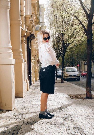 Falda de mezclilla negra con un corte relajado, combinada con una blusa blanca con bordado. Unas gafas de sol oscuras y unas bailarinas negras completan el look.
