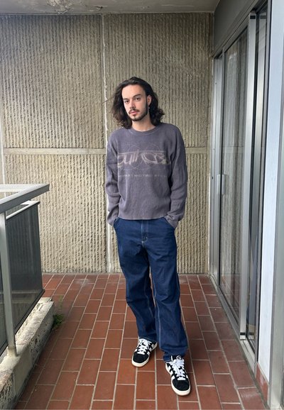Suéter gris acanalado con un logo impreso, pantalones vaqueros sueltos azules y zapatillas negras con detalles en blanco, de pie sobre una superficie de baldosas.