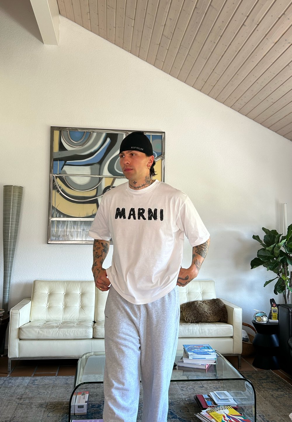 Λευκό oversized t-shirt με την εγγραφή "MARNI" σε μαύρο, συνδυασμένο με γκρι παντελόνια φόρμας. Φορεμένο μαύρο καπέλο, περιβεβλημένο από μινιμαλιστικά έπιπλα.