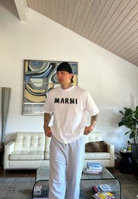 Λευκό oversized t-shirt με την εγγραφή "MARNI" σε μαύρο, συνδυασμένο με γκρι παντελόνια φόρμας. Φορεμένο μαύρο καπέλο, περιβεβλημένο από μινιμαλιστικά έπιπλα.