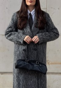 Sac à main en cuir synthétique noir avec une forme structurée, un design allongé et une texture lisse. Tenue par une personne portant un manteau gris surdimensionné.