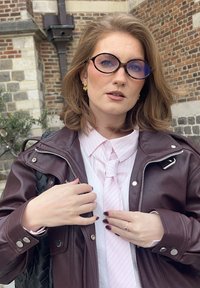 Brun läderjacka, rosa randig skjorta med slips, runda svarta glasögon med blå ton, guldfärgade örhängen, texturerad svart väska och en tegelstensbakgrund.