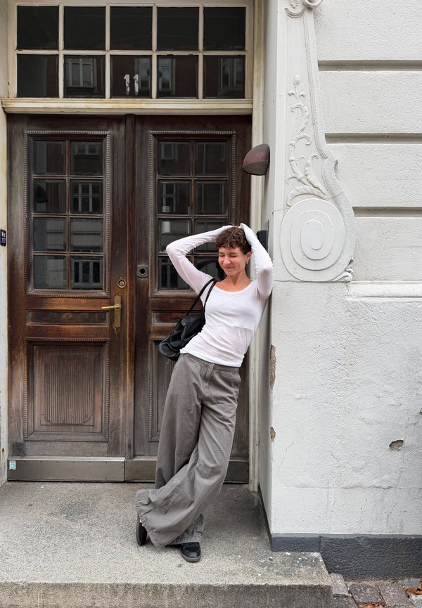 Una mujer con un top blanco y pantalones grises holgados posa con las manos en el pelo junto a una puerta ornamentada.