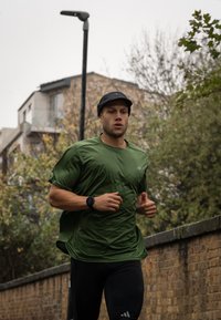 Grønn syntetisk løpetrøye med korte ermer og mørke leggings. En svart caps og klokke kompletterer antrekket. Løper på en brosteinssti.