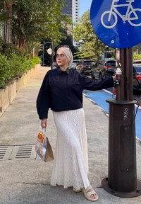 Vrouw in een zwart shirt, witte kanten rok en hoofddoek staat op de stoep met een tas, leunend op een paal met een blauw fietsbord boven haar.