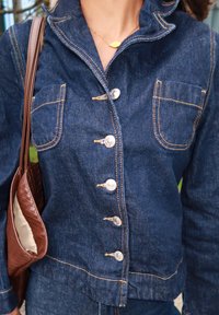 Jeansjacke in Dunkelblau mit taillierter Passform, Knopfverschluss, zwei Brusttaschen und kontrastierenden orangen Nähten. Neben ihr eine braune geflochtene Tasche.