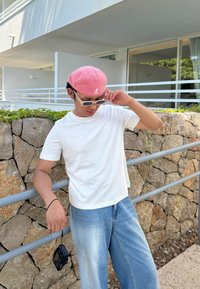 Witte T-shirt met korte mouwen, lichtblauwe jeans, roze badstof pet en een zonnebril. Staande bij een stenen muur met een camera die aan de hand hangt.
