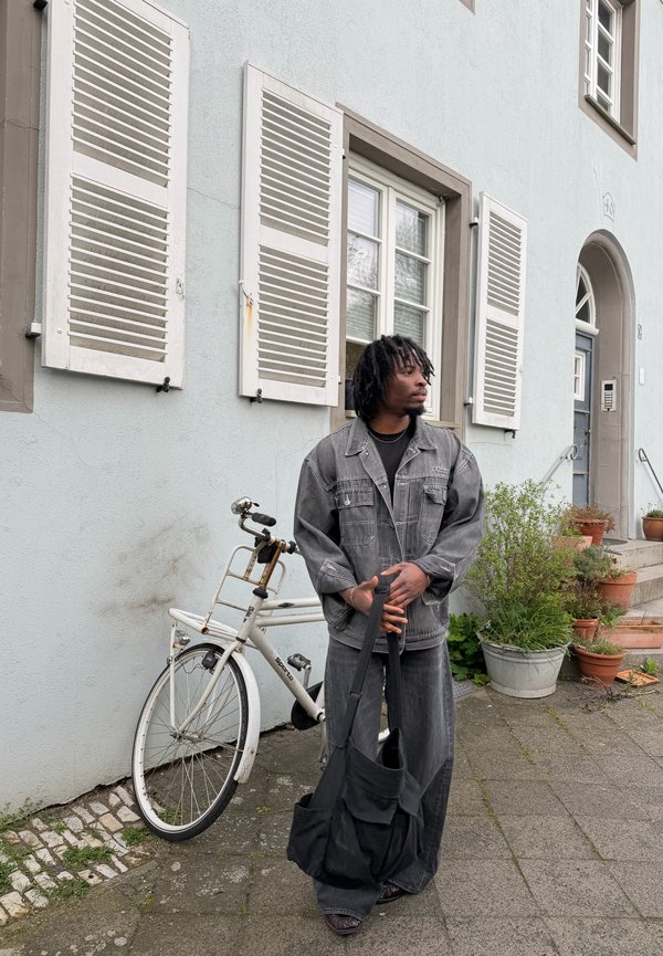 Mies harmaassa denimitakissa ja housuissa seisoo valkoisen polkupyörän vieressä rakennuksen ulkopuolella, jossa on valkoiset luukkulasi-ikkunat ja ruukkukasveja.
