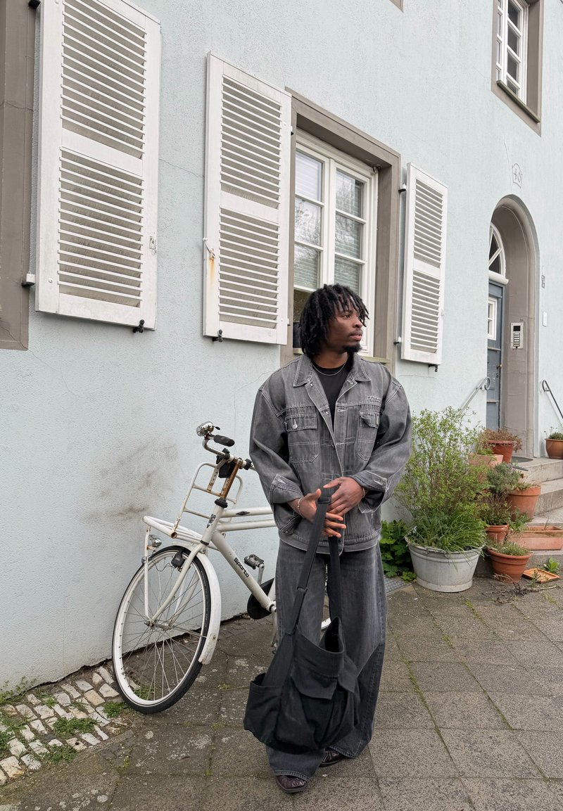 Homme en veste et pantalon en jean gris se tient près d'un vélo blanc devant un bâtiment avec des fenêtres blanches à volets et des plantes en pot.