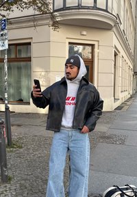 Veste courte en cuir noir, sweat à capuche gris avec un graphisme rouge, jean large bleu clair. Debout sur un trottoir, tenant un smartphone.