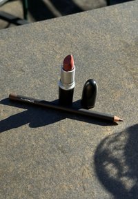 Rouge à lèvres MAC dans un tube argenté avec une pointe arrondie, teinte rose, à côté d'un crayon à lèvres noir avec une pointe taillée.