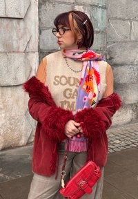 Άτομο που φοράει κόκκινο παλτό από ψεύτικη γούνα, πολύχρωμο φλοράλ κασκόλ, γυαλιά και κρατάει μια κόκκινη τσάντα, στέκεται μπροστά σε γκρι πέτρινο τοίχο.