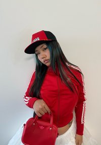 Rdeča zip-up športna jakna z belimi črtami, v kombinaciji s črno-rdečo kapo. Majhna rdeča torbica dopolnjuje videz. Vidna bela tila krila.