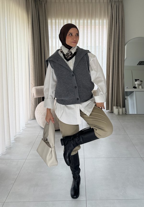 Une femme portant un hijab foncé pose à l’intérieur. Elle tient un sac à main beige et porte un gilet gris, une chemise blanche, un pantalon beige et des bottes noires jusqu’aux genoux.