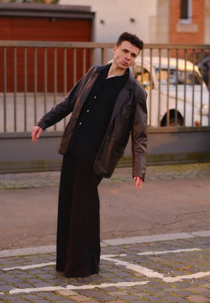 Chaqueta de cuero negra sobre una camisa negra, combinada con pantalones anchos negros. El modelo está en una pose inclinada contra una superficie de piedra.