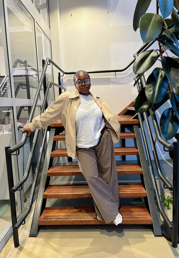 Chaqueta oversized beige sobre una camiseta blanca, combinada con pantalones de pierna ancha beige. De pie en unas escaleras de madera con barandillas de metal negro y vegetación.