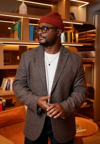 Blazer en pied-de-poule marron porté sur une chemise blanche, avec un bonnet rouge tricoté. Collier en argent avec pendentif, encadré par des étagères en bois et des livres.