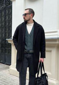 Fekete gyapjú nagykabát zöld gombos kardigán, krémszínű pulóver és fekete farmer felett. Fekete táskát tart a kezében. A háttérben díszes ajtó látható.