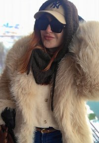 Femme portant un manteau en fourrure beige, une écharpe noire, une casquette beige, de grandes lunettes de soleil noires et un jean bleu, posant en extérieur par temps froid.