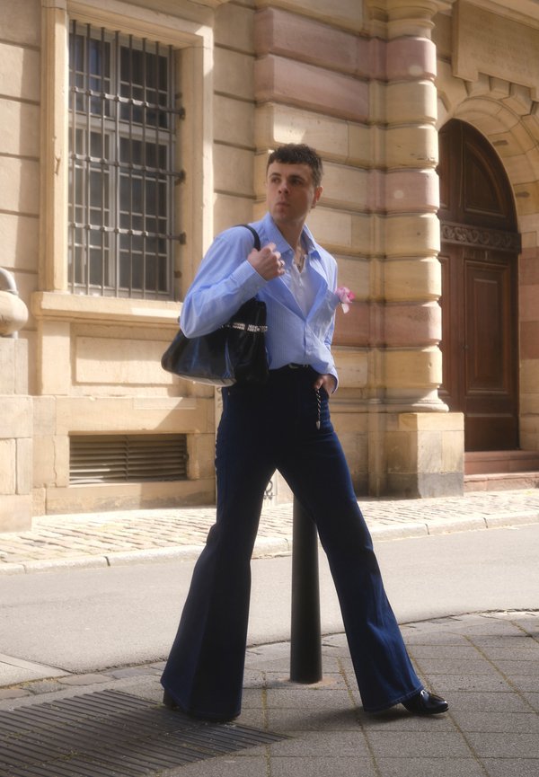 Uomo con camicia blu abbottonata e pantaloni scuri a zampa che tiene una borsa nera, in piedi su una strada cittadina vicino a un edificio di pietra con grandi finestre e porta in legno.