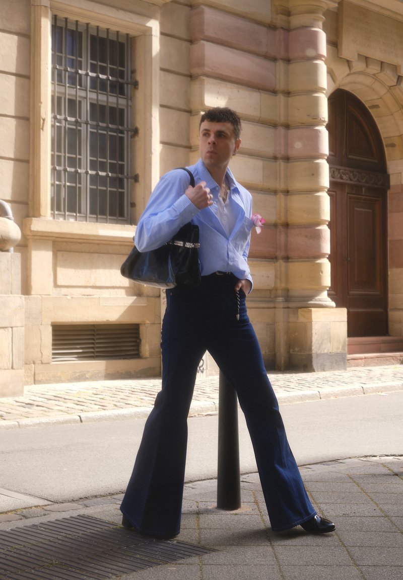 Homme en chemise bleue boutonnée et pantalon évasé foncé tenant un sac noir, debout dans une rue de la ville près d'un bâtiment en pierre avec de grandes fenêtres et une porte en bois.