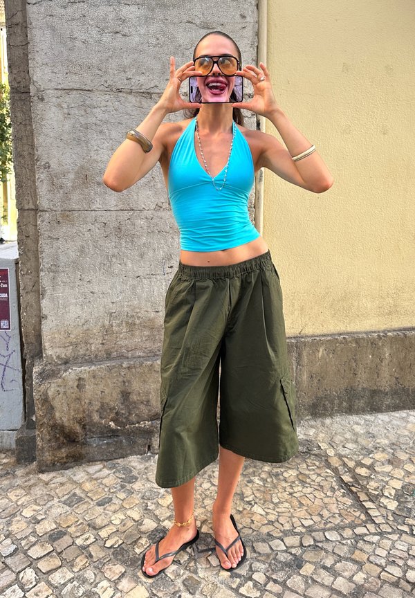 Femme en haut bleu et pantalon vert tient un téléphone affichant sa bouche souriante sur son visage, portant de grandes lunettes de soleil, debout dans une rue pavée.