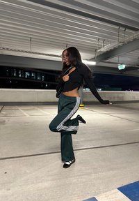 Schwarzes geripptes Crop-Top, grüne Jogginghose mit weißen Streifen und schwarze Schuhe. Innenparkhaus mit Deckenlichtern und Beschilderung.