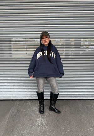 Sudadera marina con "BROOKLYN" en letras crema, combinada con jeans grises y botas negras hasta la rodilla, contra un fondo de puerta enrollable metálica.