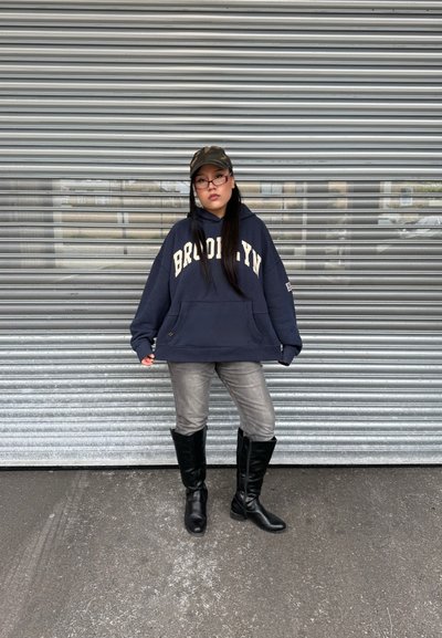 Sudadera marina con "BROOKLYN" en letras crema, combinada con jeans grises y botas negras hasta la rodilla, contra un fondo de puerta enrollable metálica.