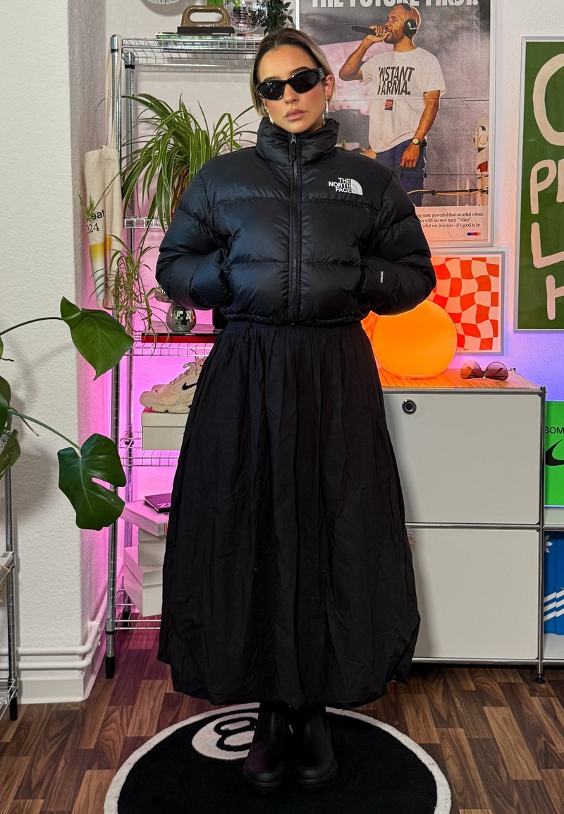 Casaco puffer preto com costuras verticais, gola alta e logótipo. Saia preta fluida com cintura franzida. Botas pretas de tornozelo completam o look.