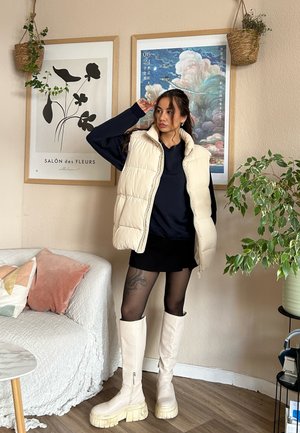 Chaleco de crema acolchado sobre una blusa navy, combinado con una falda negra. Botas de caña alta en un beige claro con suelas gruesas. La decoración incluye plantas en macetas y arte.