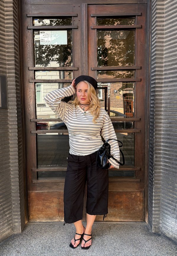 Donna con basco nero, maglia a righe a maniche lunghe, pantaloni neri larghi e sandali, in piedi accanto a una porta in legno tenendo una borsa nera.