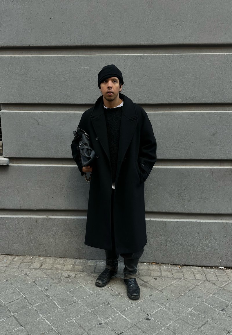 Black overcoat with large lapels, worn over a textured black jumper. Holding a black bag, paired with black boots, standing on a cobblestone street.