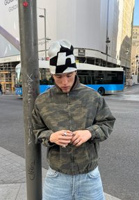 Jaunuolis kamufliažinėje striukėje ir juodai baltame kepurytėje žiūri žemyn, reguliuodamas žiedus, stovėdamas prie gatvės žibinto su mėlynu autobusu ir miesto fonu.