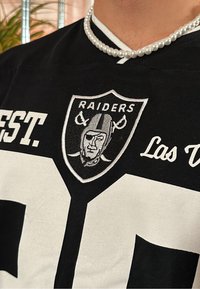 Črna športna majica z belim številom '21' in vezenim logotipom Raiders z likom v čeladi. Ovratnik krasijo biserni dodatki.