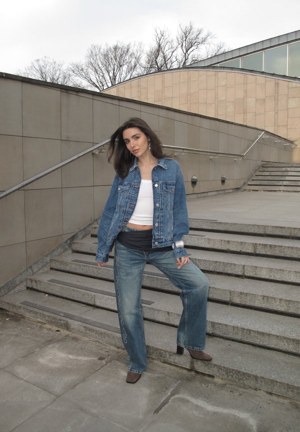 Tânără într-o jachetă din denim și blugi, sprijinită de scările urbane, cu copaci dezgoliți și o clădire modernă în fundal.