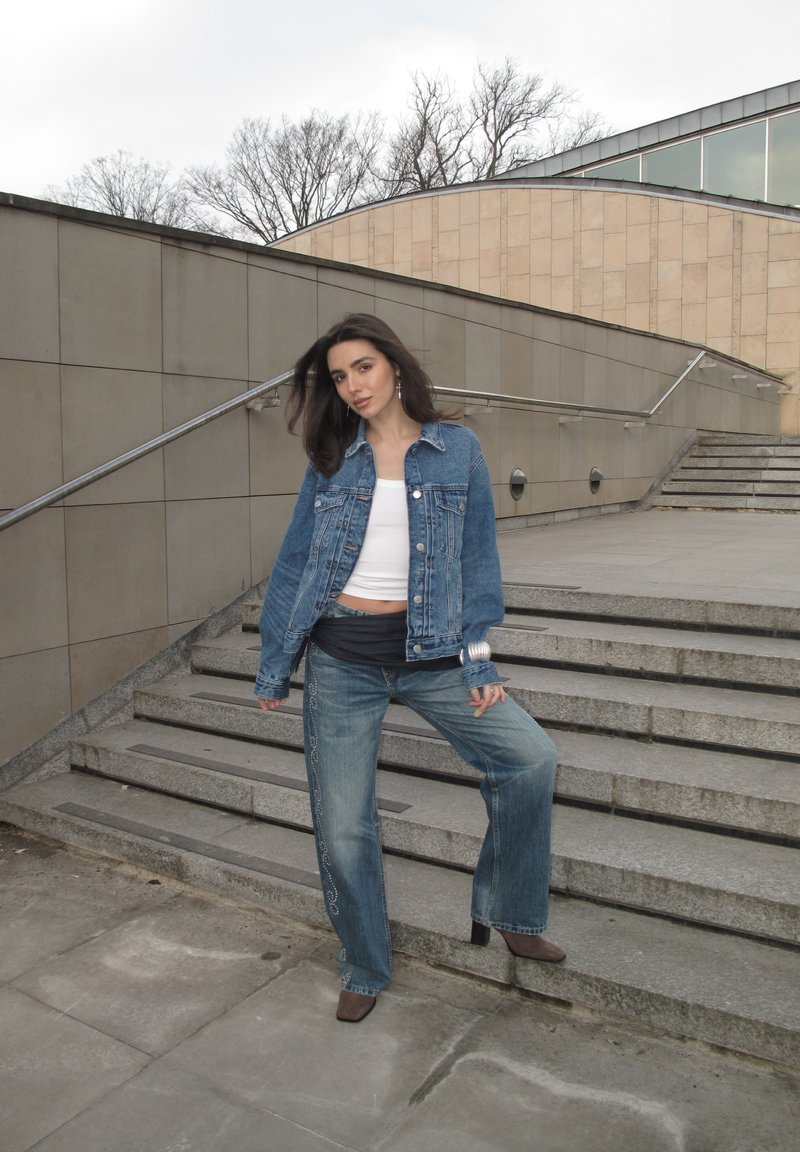 Mlada žena u traper jakni i trapericama naslonjena na gradska stepenice, u pozadini goli drveće i moderna zgrada.