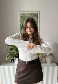 Hvit ribbestrikket genser med lange ermer og utsvingte mansjetter, kombinert med en mørkebrun pinstripet skjørt og svart belte. I bakgrunnen er det en plante og en lampe.